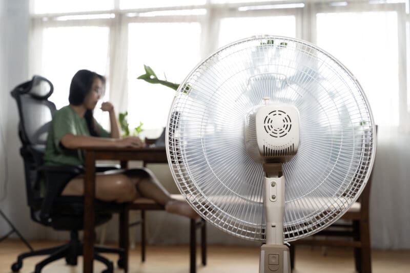 Standing electric fan in a home office with a person seated at a desk in the background
