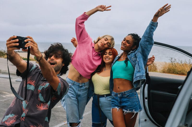 Four friends celebrating and taking a selfie beside a car at a coastal parking area