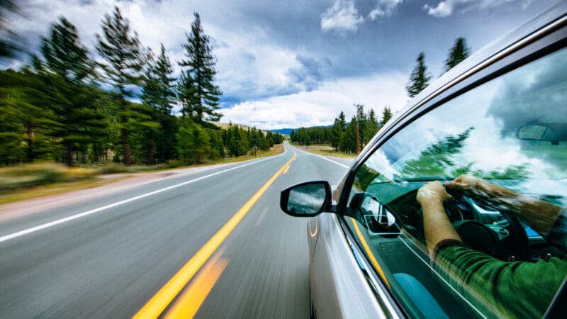 Close-up side view of a car driving at speed on a tree-lined highway
